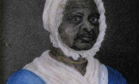 A framed portrait of a Black woman wearing a blue dress with drawstrings at the neck and waist, a white fichu tucked into her dress at the neck, a white cap, and a gold beaded necklace.